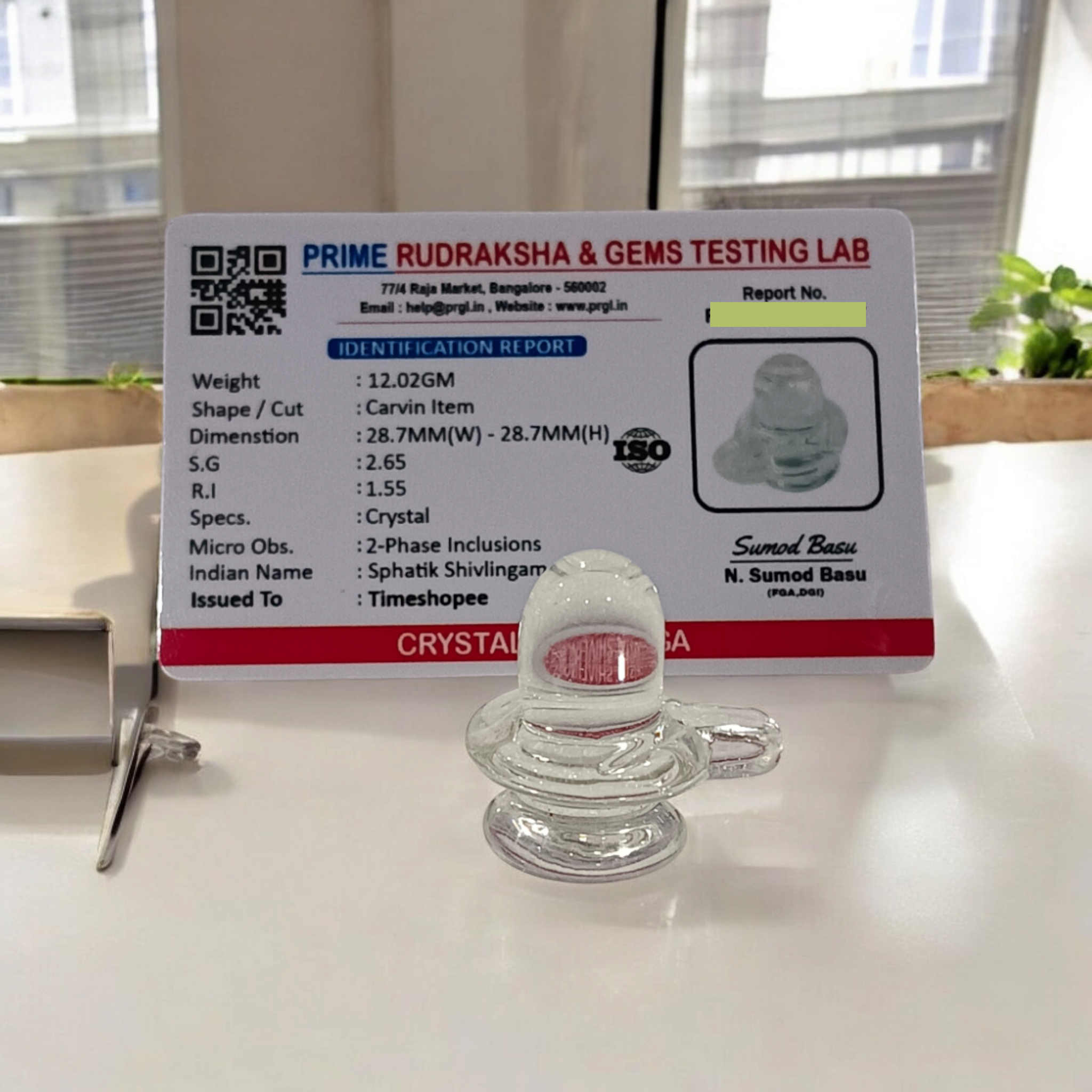 A clear, polished Sphatik (Crystal) Shivling placed on a small round base, intricately designed with fine details. The Shivling radiates purity and divine energy, symbolizing Lord Shiva. Ideal for worship, this lab-certified Sphatik Shivling from Timeshopee adds a spiritual touch to your home or temple setting.