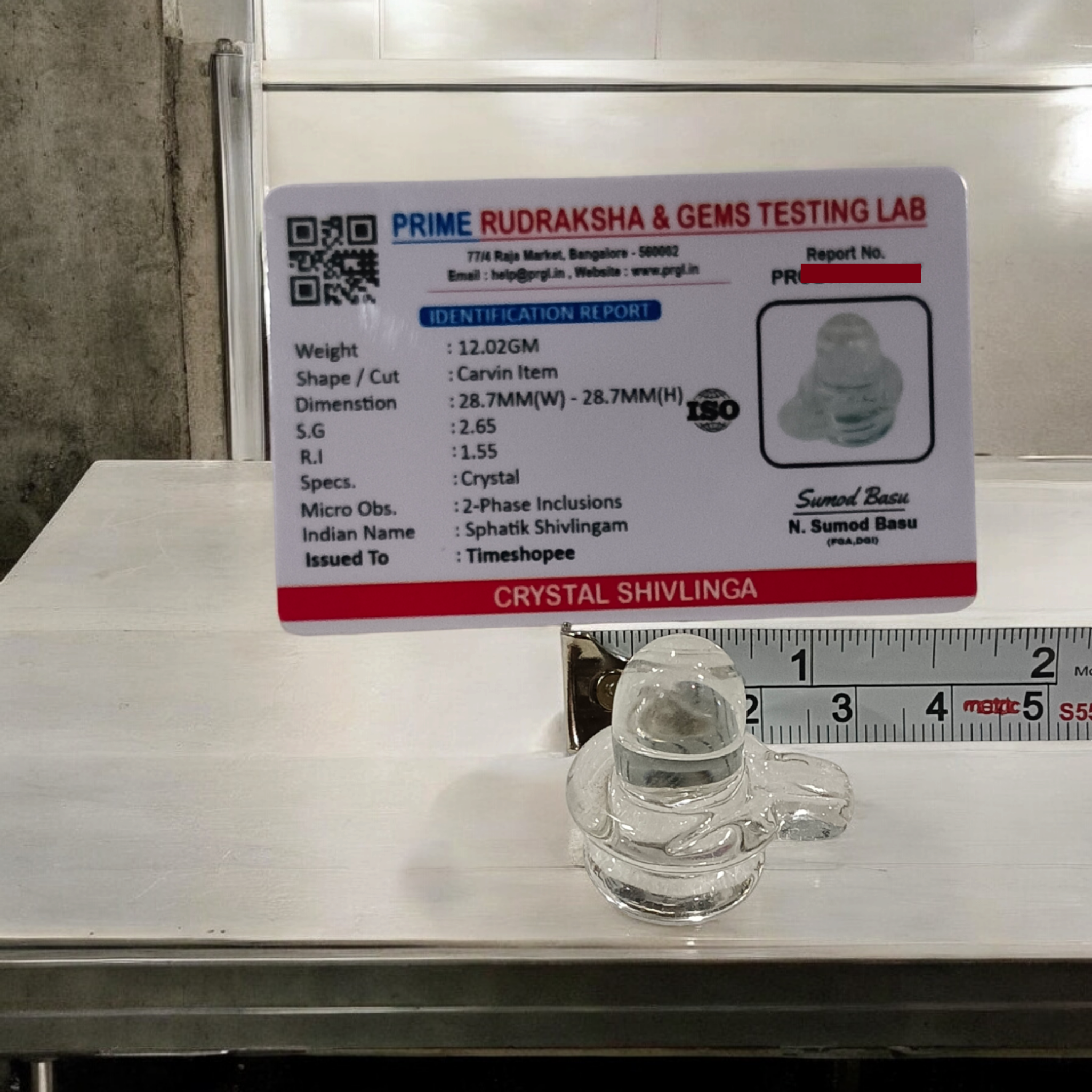 A clear, polished Sphatik (Crystal) Shivling placed on a small round base, intricately designed with fine details. The Shivling radiates purity and divine energy, symbolizing Lord Shiva. Ideal for worship, this lab-certified Sphatik Shivling from Timeshopee adds a spiritual touch to your home or temple setting.