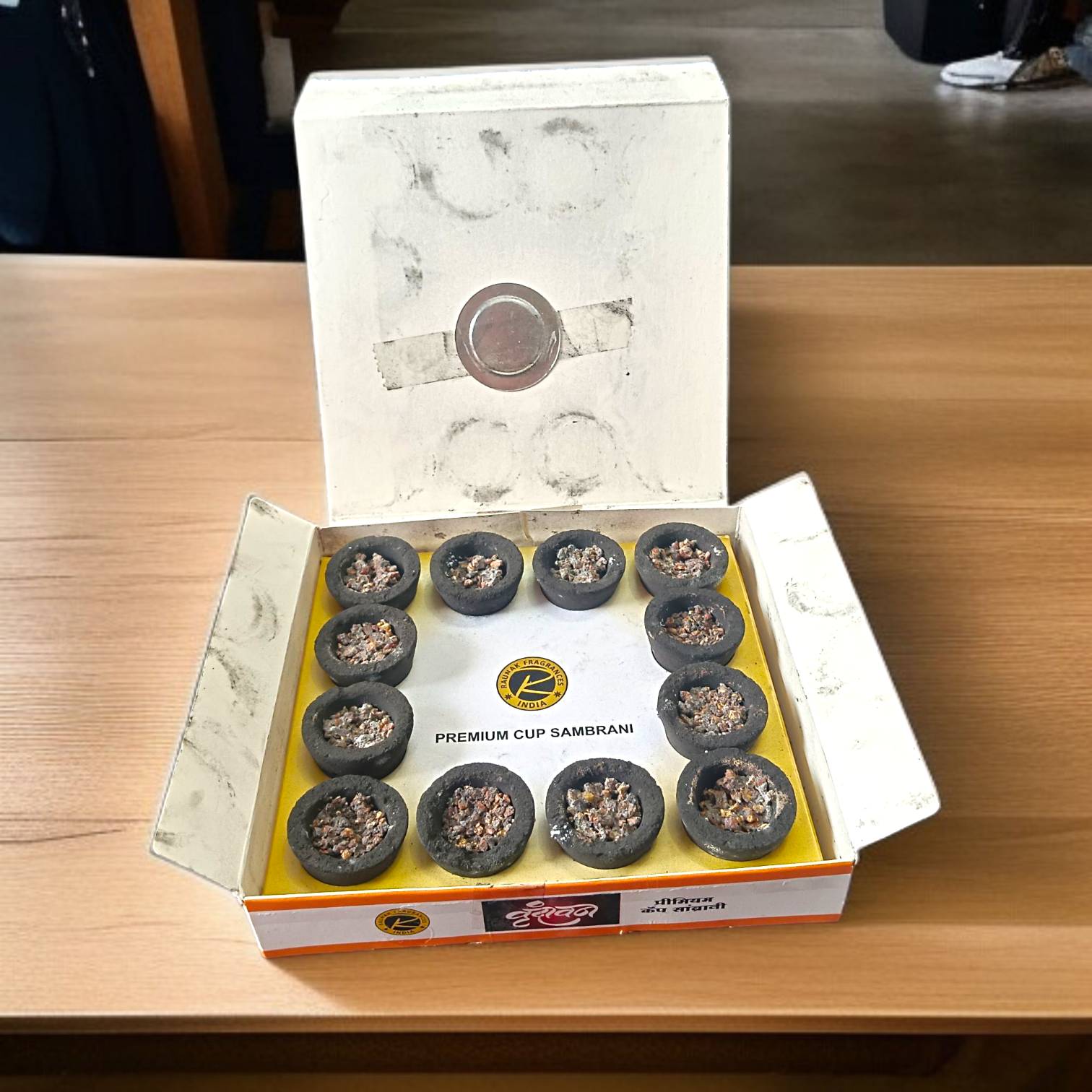 A box of Premium Cup Sambrani containing 12 natural dhoop cups arranged neatly, with herbal ingredients in the background