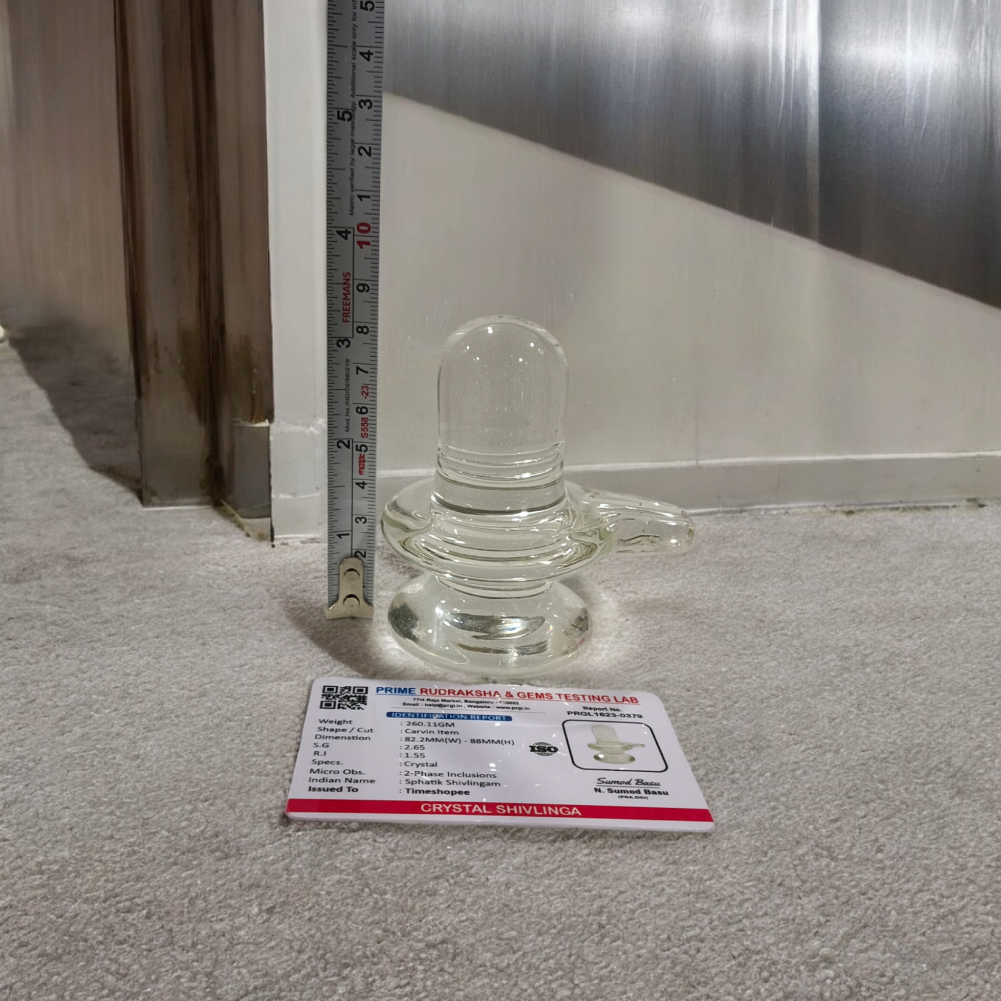 A clear, polished Sphatik (Crystal) Shivling placed on a small round base, intricately designed with fine details. The Shivling radiates purity and divine energy, symbolizing Lord Shiva. Ideal for worship, this lab-certified Sphatik Shivling from Timeshopee adds a spiritual touch to your home or temple setting.