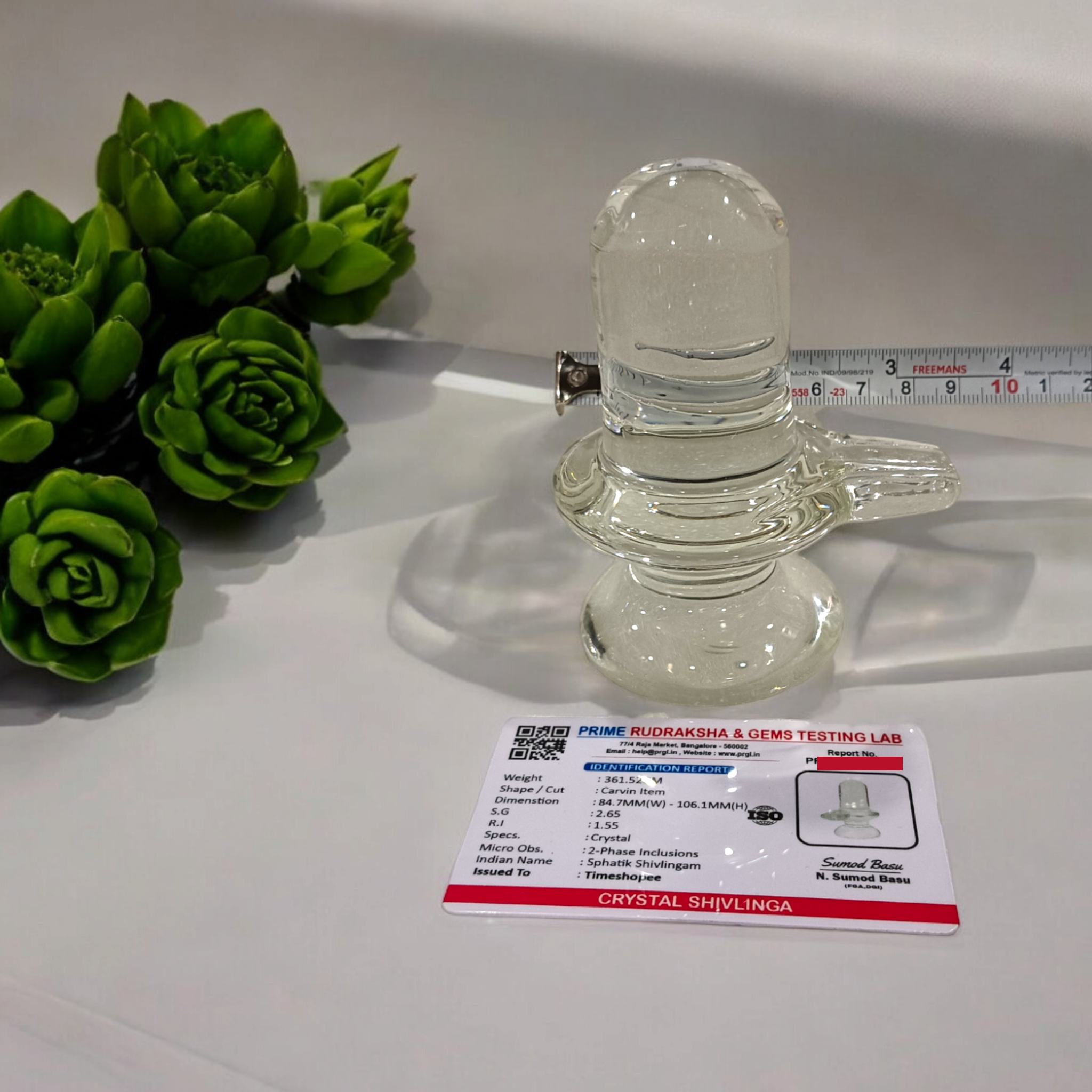 A clear, polished Sphatik (Crystal) Shivling placed on a small round base, intricately designed with fine details. The Shivling radiates purity and divine energy, symbolizing Lord Shiva. Ideal for worship, this lab-certified Sphatik Shivling from Timeshopee adds a spiritual touch to your home or temple setting.