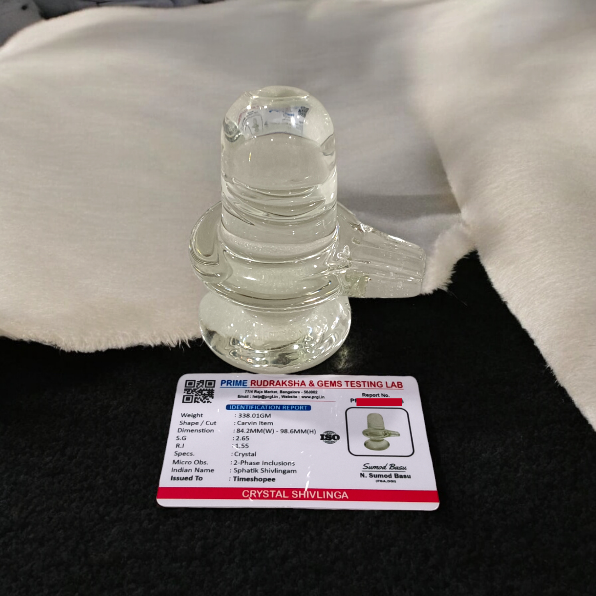 A clear, polished Sphatik (Crystal) Shivling placed on a small round base, intricately designed with fine details. The Shivling radiates purity and divine energy, symbolizing Lord Shiva. Ideal for worship, this lab-certified Sphatik Shivling from Timeshopee adds a spiritual touch to your home or temple setting.