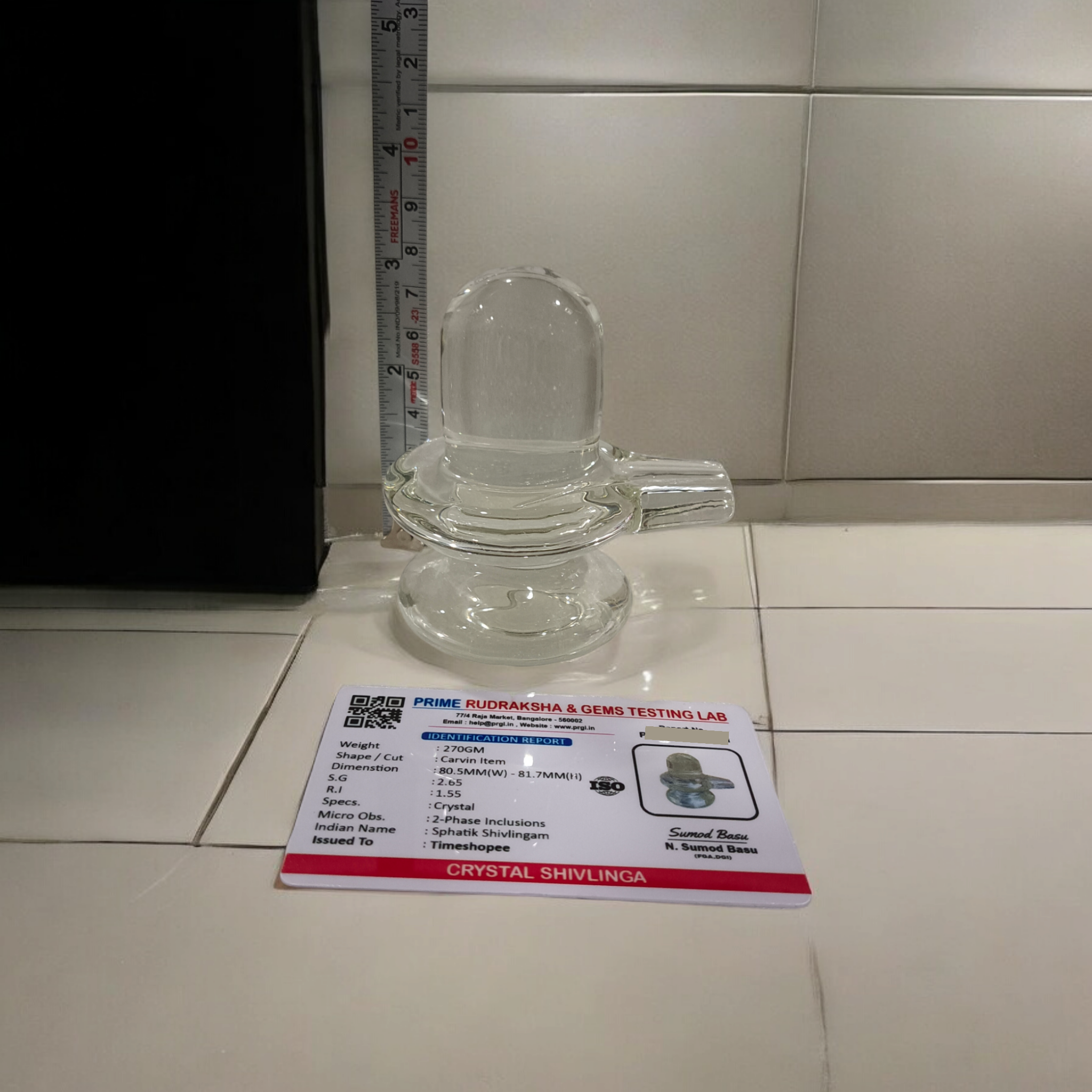 A clear, polished Sphatik (Crystal) Shivling placed on a small round base, intricately designed with fine details. The Shivling radiates purity and divine energy, symbolizing Lord Shiva. Ideal for worship, this lab-certified Sphatik Shivling from Timeshopee adds a spiritual touch to your home or temple setting.