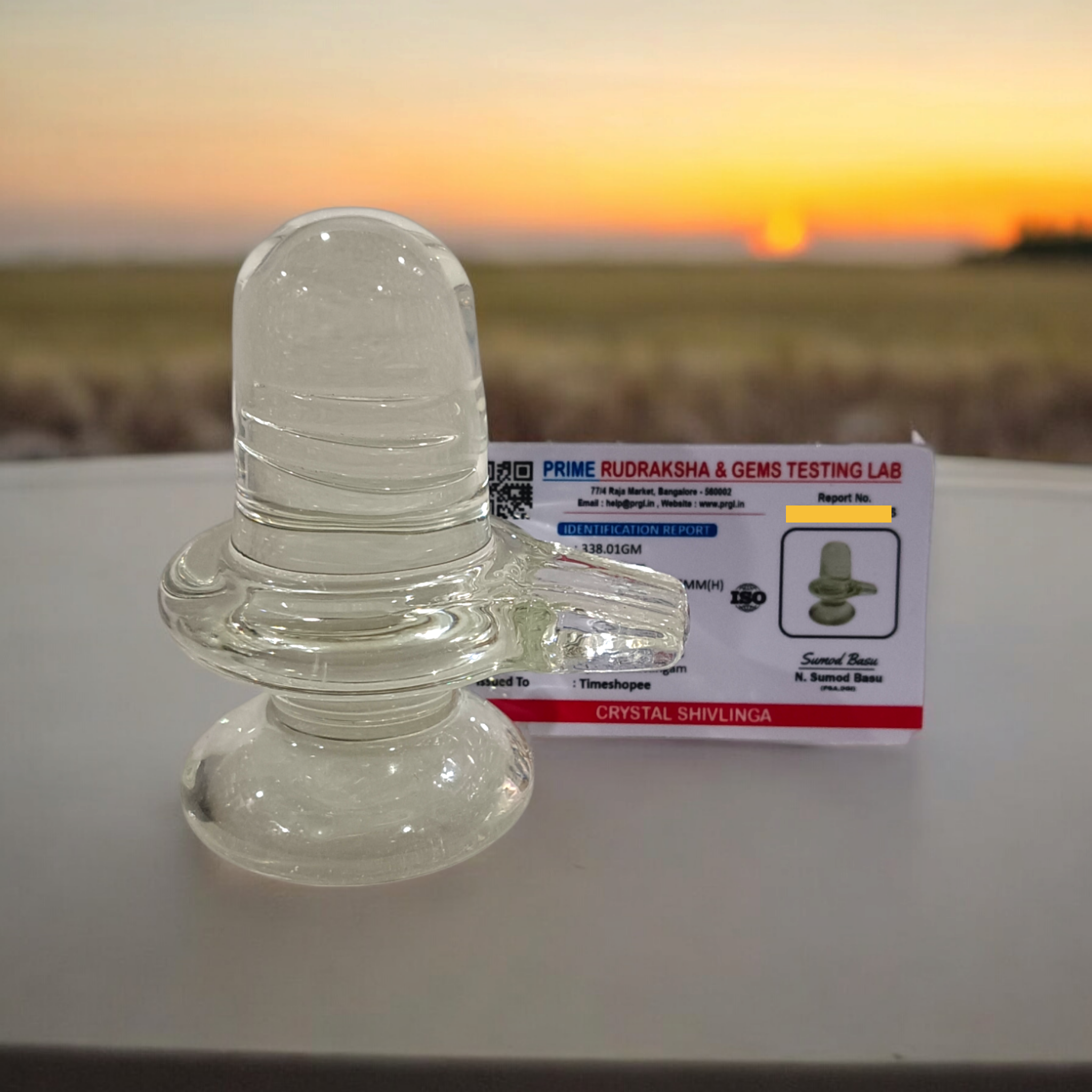 A clear, polished Sphatik (Crystal) Shivling placed on a small round base, intricately designed with fine details. The Shivling radiates purity and divine energy, symbolizing Lord Shiva. Ideal for worship, this lab-certified Sphatik Shivling from Timeshopee adds a spiritual touch to your home or temple setting.