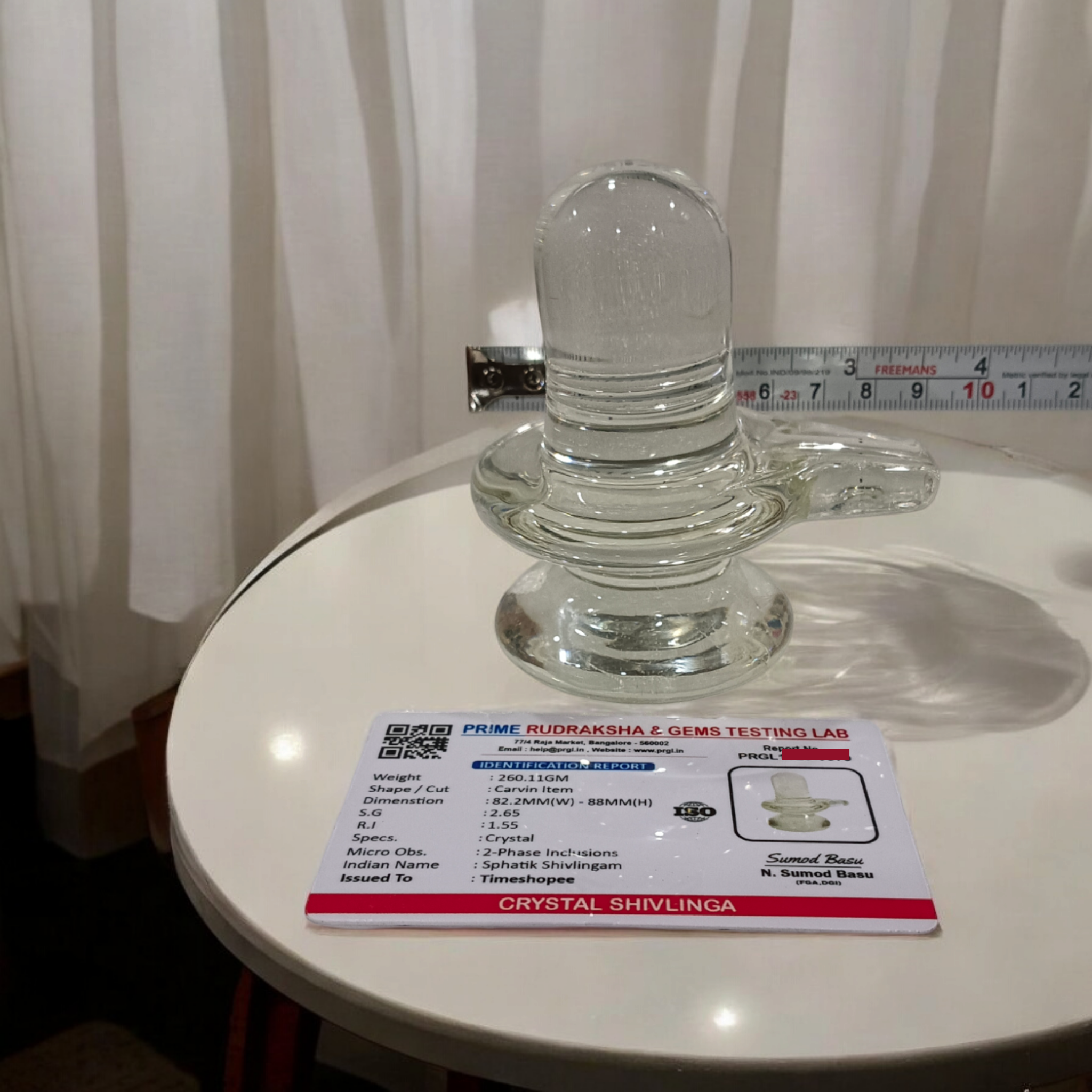 A clear, polished Sphatik (Crystal) Shivling placed on a small round base, intricately designed with fine details. The Shivling radiates purity and divine energy, symbolizing Lord Shiva. Ideal for worship, this lab-certified Sphatik Shivling from Timeshopee adds a spiritual touch to your home or temple setting.