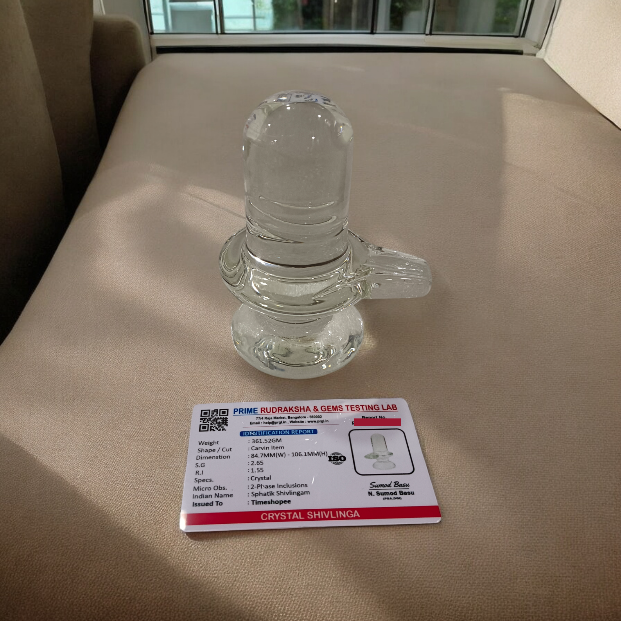 A clear, polished Sphatik (Crystal) Shivling placed on a small round base, intricately designed with fine details. The Shivling radiates purity and divine energy, symbolizing Lord Shiva. Ideal for worship, this lab-certified Sphatik Shivling from Timeshopee adds a spiritual touch to your home or temple setting.