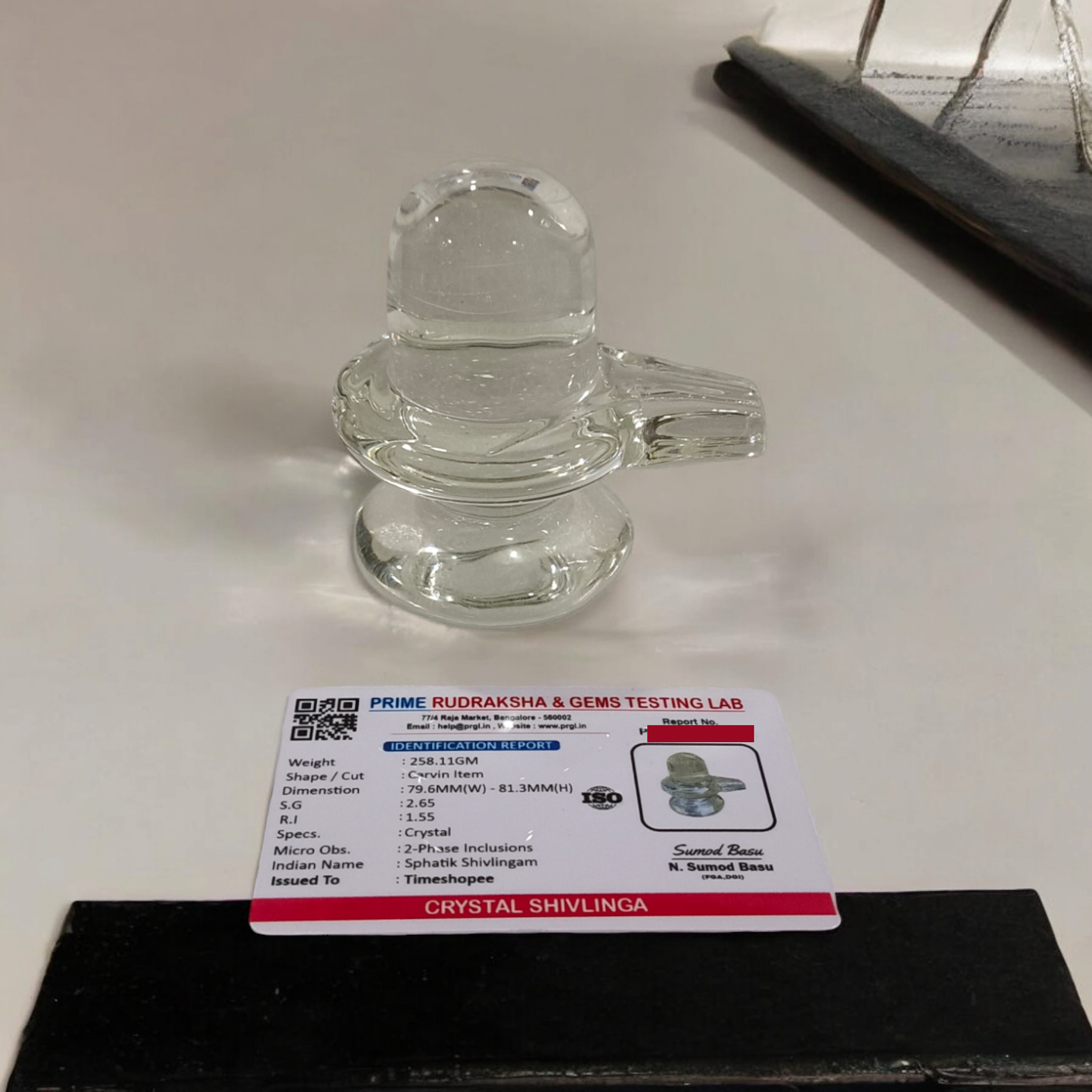 A clear, polished Sphatik (Crystal) Shivling placed on a small round base, intricately designed with fine details. The Shivling radiates purity and divine energy, symbolizing Lord Shiva. Ideal for worship, this lab-certified Sphatik Shivling from Timeshopee adds a spiritual touch to your home or temple setting.