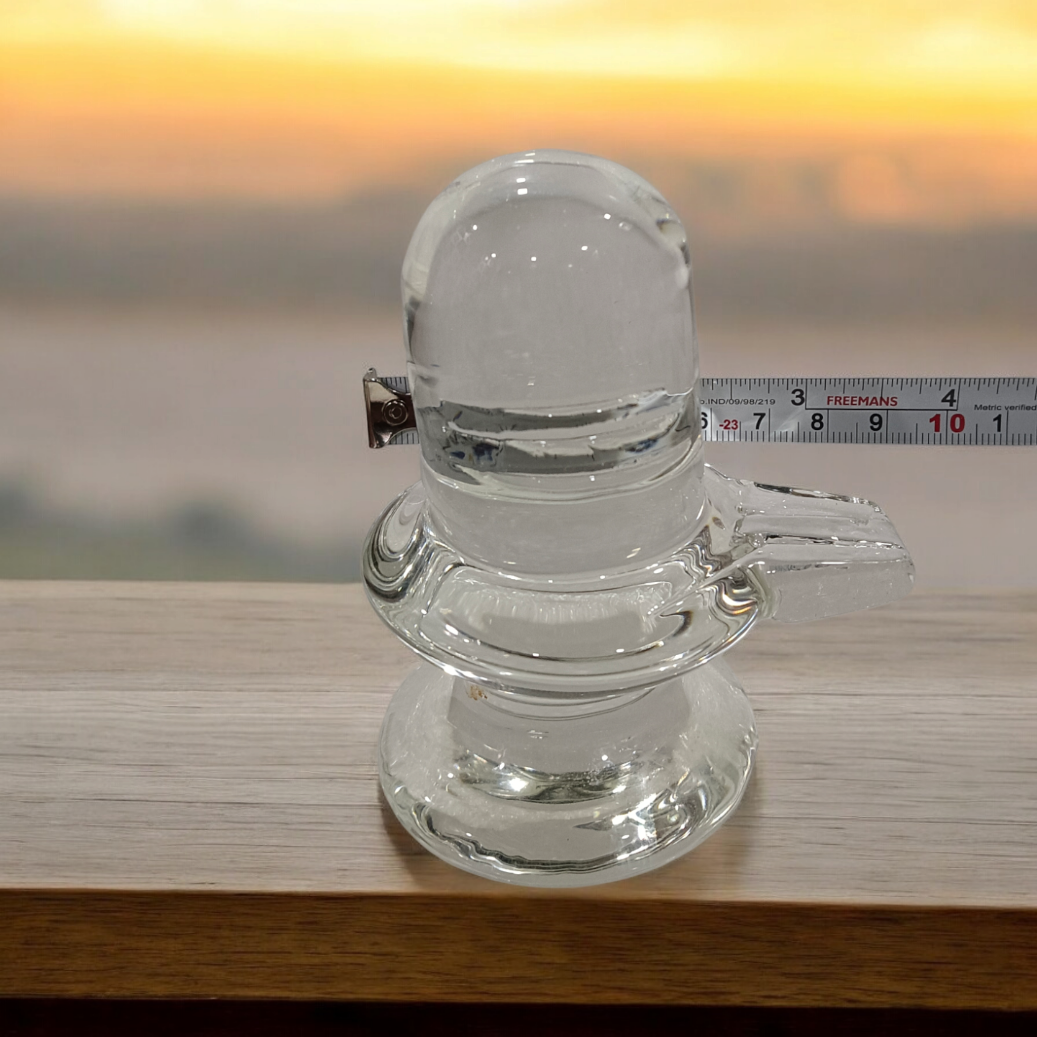 A clear, polished Sphatik (Crystal) Shivling placed on a small round base, intricately designed with fine details. The Shivling radiates purity and divine energy, symbolizing Lord Shiva. Ideal for worship, this lab-certified Sphatik Shivling from Timeshopee adds a spiritual touch to your home or temple setting.