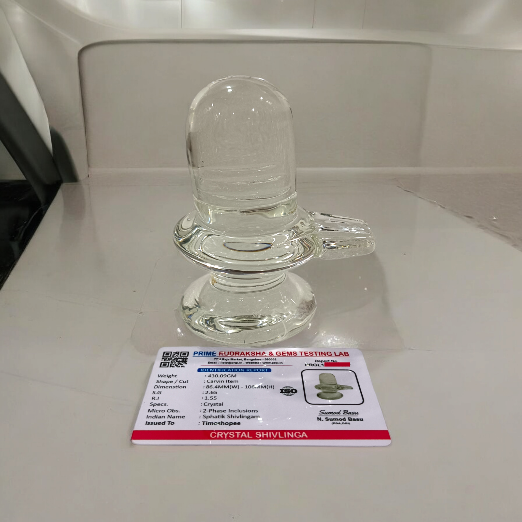 A clear, polished Sphatik (Crystal) Shivling placed on a small round base, intricately designed with fine details. The Shivling radiates purity and divine energy, symbolizing Lord Shiva. Ideal for worship, this lab-certified Sphatik Shivling from Timeshopee adds a spiritual touch to your home or temple setting.