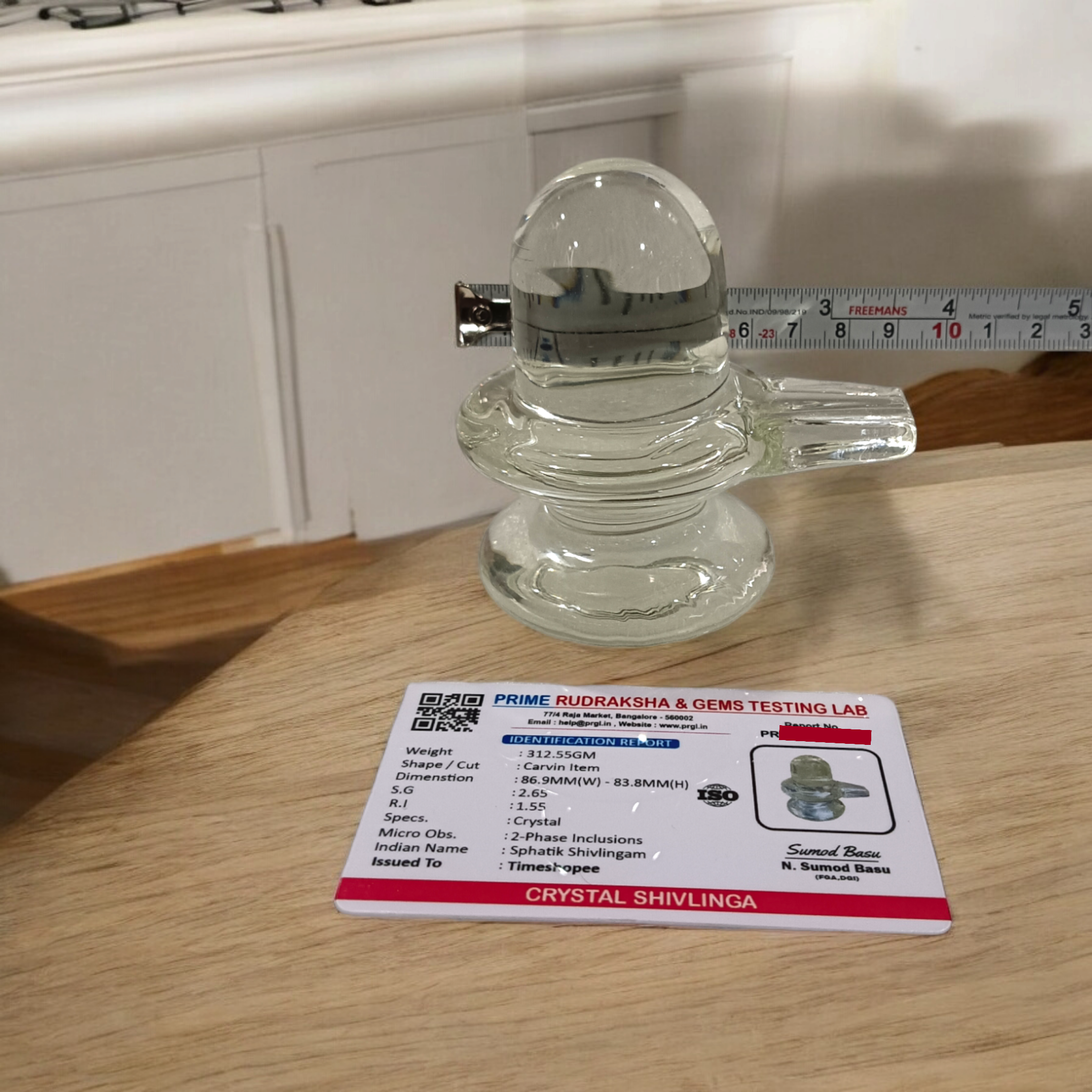 A clear, polished Sphatik (Crystal) Shivling placed on a small round base, intricately designed with fine details. The Shivling radiates purity and divine energy, symbolizing Lord Shiva. Ideal for worship, this lab-certified Sphatik Shivling from Timeshopee adds a spiritual touch to your home or temple setting.