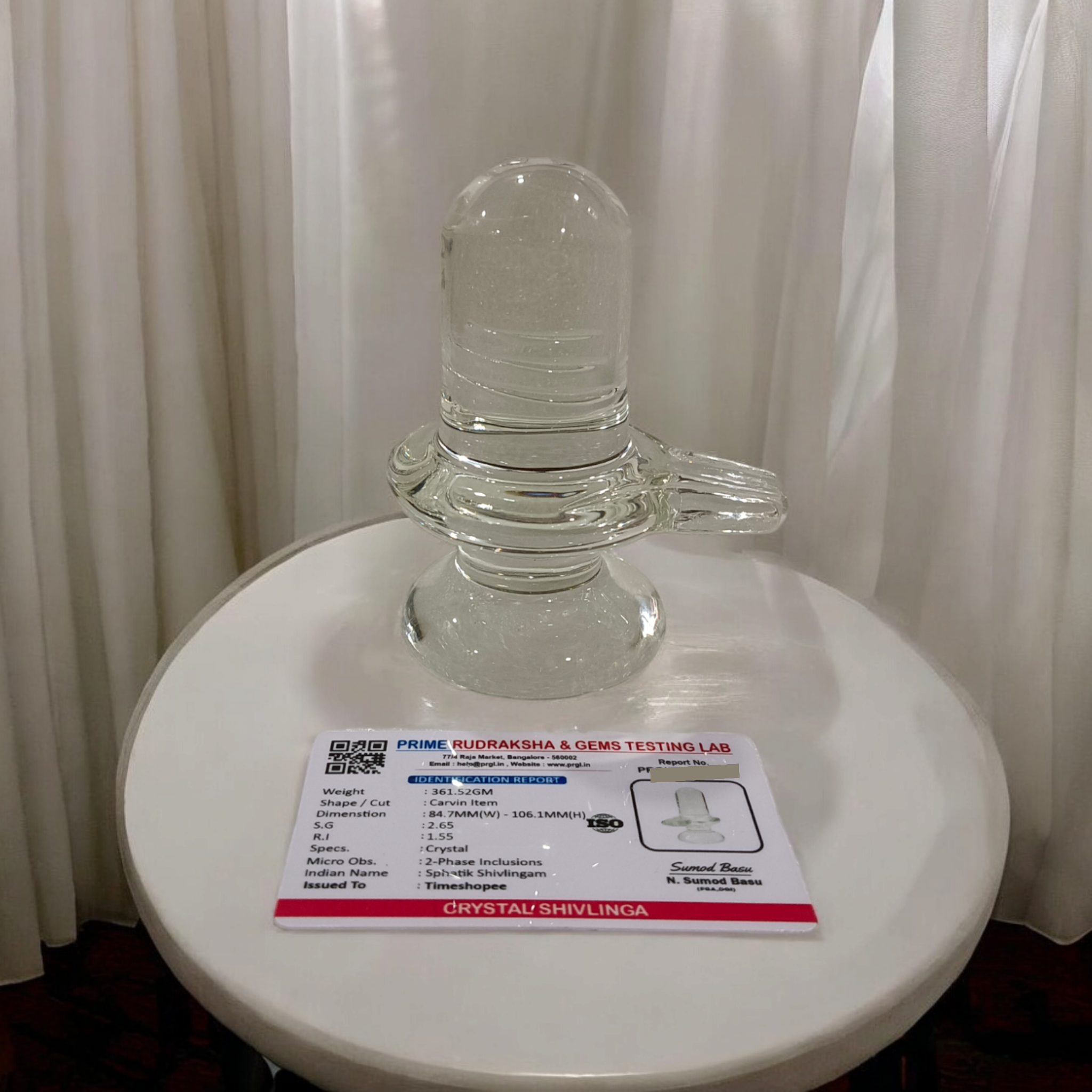 A clear, polished Sphatik (Crystal) Shivling placed on a small round base, intricately designed with fine details. The Shivling radiates purity and divine energy, symbolizing Lord Shiva. Ideal for worship, this lab-certified Sphatik Shivling from Timeshopee adds a spiritual touch to your home or temple setting.