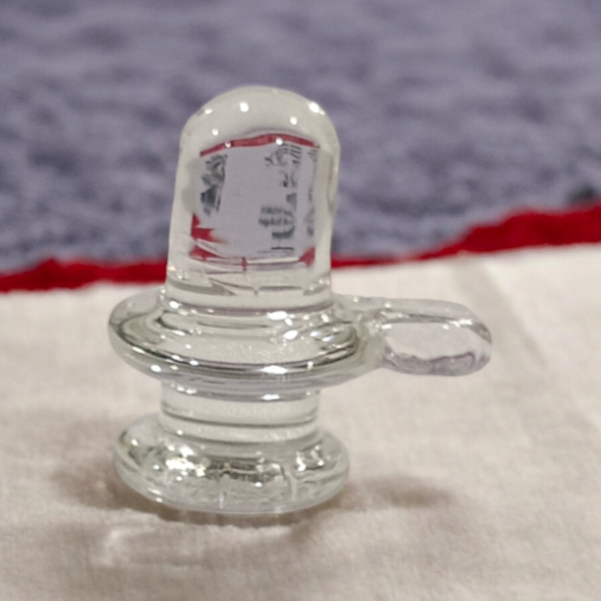 A clear, polished Sphatik (Crystal) Shivling placed on a small round base, intricately designed with fine details. The Shivling radiates purity and divine energy, symbolizing Lord Shiva. Ideal for worship, this lab-certified Sphatik Shivling from Timeshopee adds a spiritual touch to your home or temple setting.