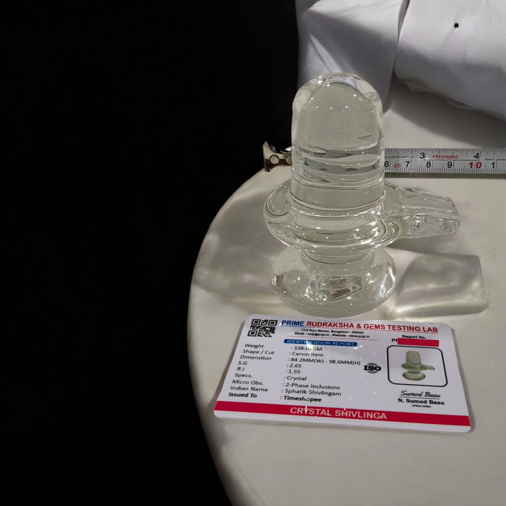 A clear, polished Sphatik (Crystal) Shivling placed on a small round base, intricately designed with fine details. The Shivling radiates purity and divine energy, symbolizing Lord Shiva. Ideal for worship, this lab-certified Sphatik Shivling from Timeshopee adds a spiritual touch to your home or temple setting.