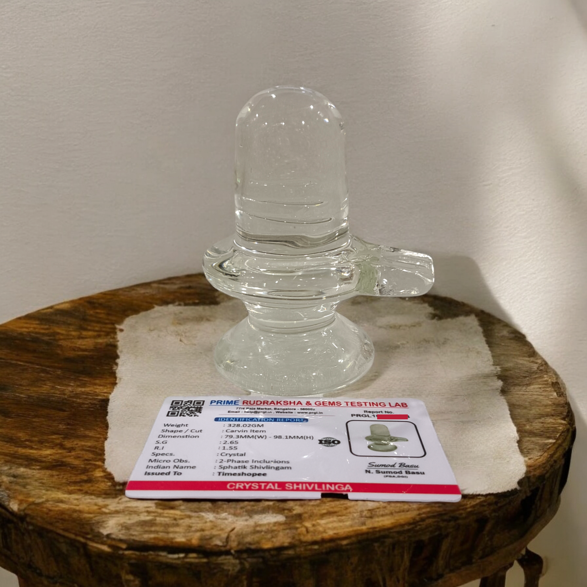 A clear, polished Sphatik (Crystal) Shivling placed on a small round base, intricately designed with fine details. The Shivling radiates purity and divine energy, symbolizing Lord Shiva. Ideal for worship, this lab-certified Sphatik Shivling from Timeshopee adds a spiritual touch to your home or temple setting.