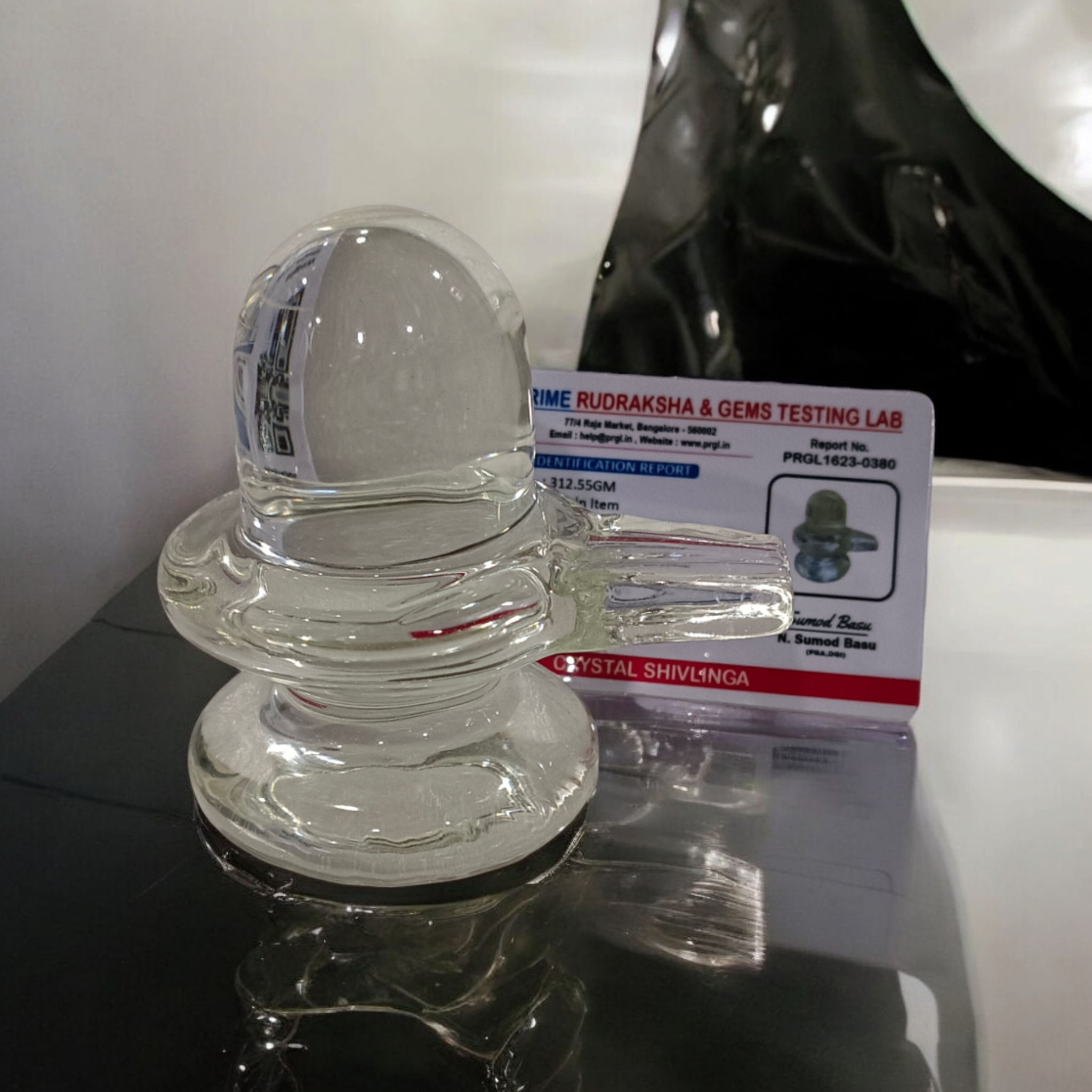 A clear, polished Sphatik (Crystal) Shivling placed on a small round base, intricately designed with fine details. The Shivling radiates purity and divine energy, symbolizing Lord Shiva. Ideal for worship, this lab-certified Sphatik Shivling from Timeshopee adds a spiritual touch to your home or temple setting.