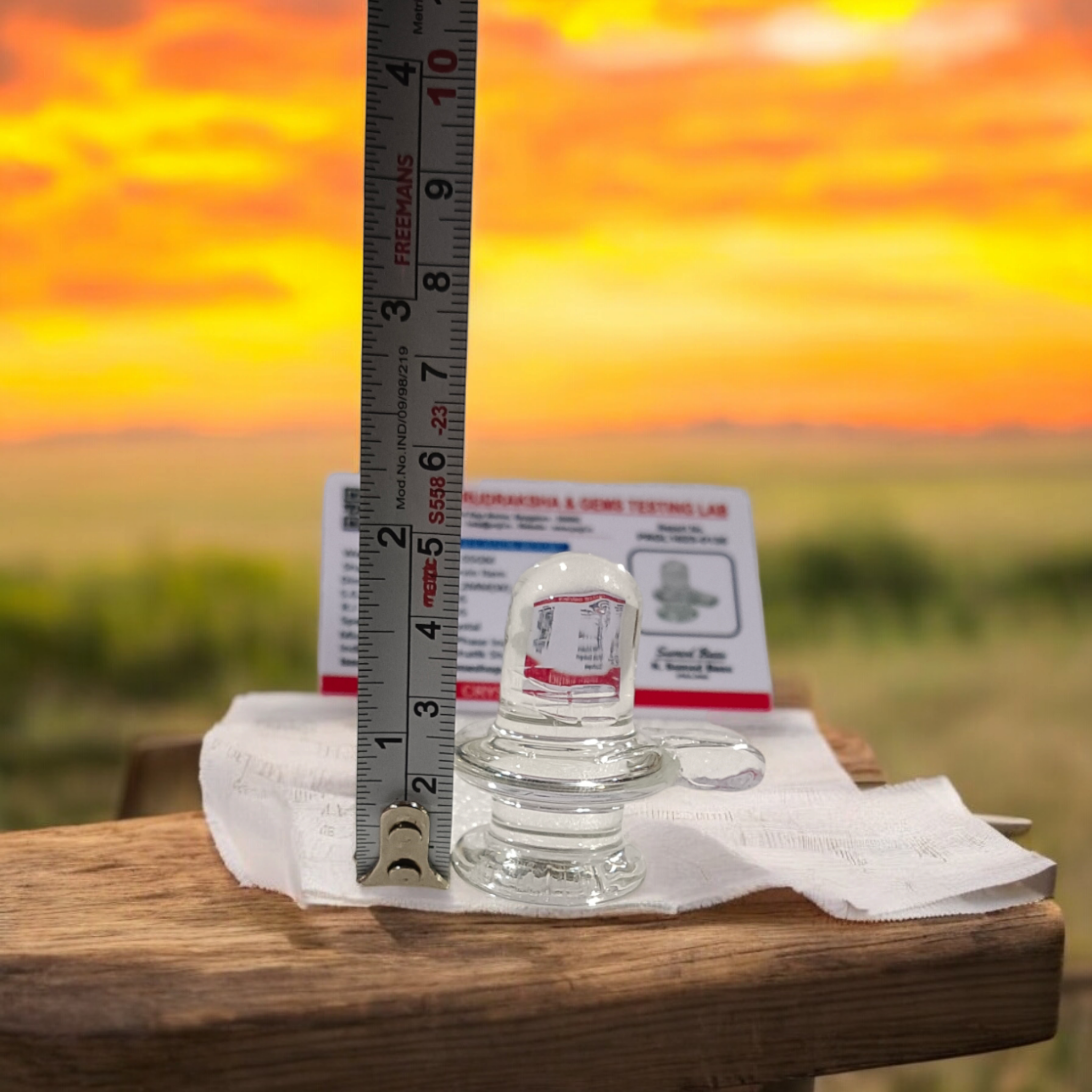 A clear, polished Sphatik (Crystal) Shivling placed on a small round base, intricately designed with fine details. The Shivling radiates purity and divine energy, symbolizing Lord Shiva. Ideal for worship, this lab-certified Sphatik Shivling from Timeshopee adds a spiritual touch to your home or temple setting.