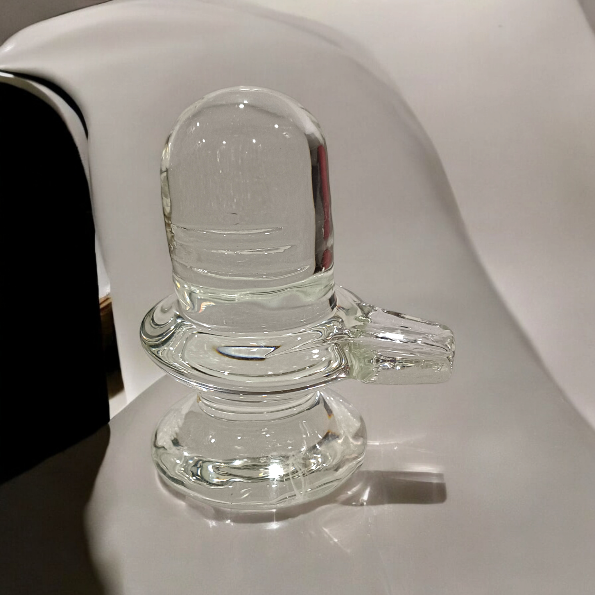 A clear, polished Sphatik (Crystal) Shivling placed on a small round base, intricately designed with fine details. The Shivling radiates purity and divine energy, symbolizing Lord Shiva. Ideal for worship, this lab-certified Sphatik Shivling from Timeshopee adds a spiritual touch to your home or temple setting.
