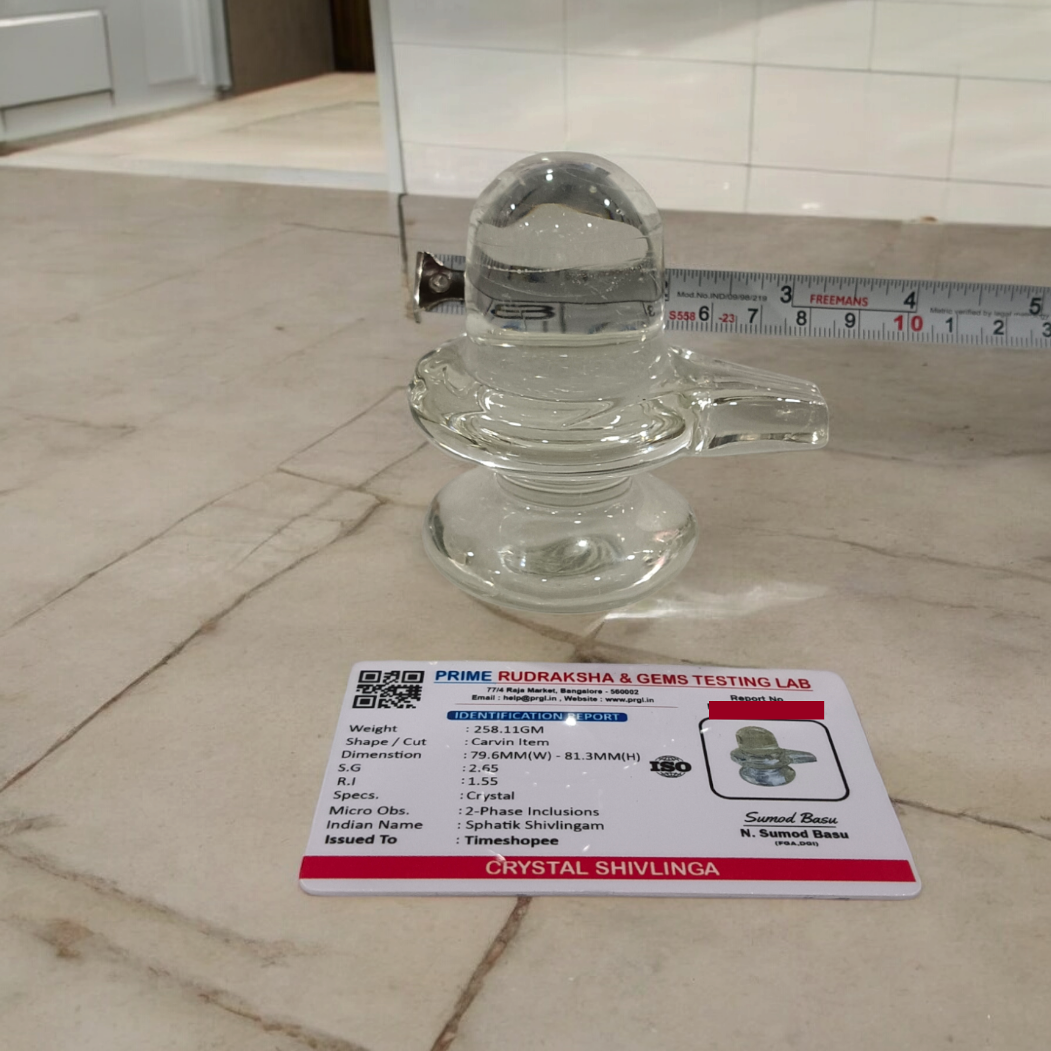 A clear, polished Sphatik (Crystal) Shivling placed on a small round base, intricately designed with fine details. The Shivling radiates purity and divine energy, symbolizing Lord Shiva. Ideal for worship, this lab-certified Sphatik Shivling from Timeshopee adds a spiritual touch to your home or temple setting.