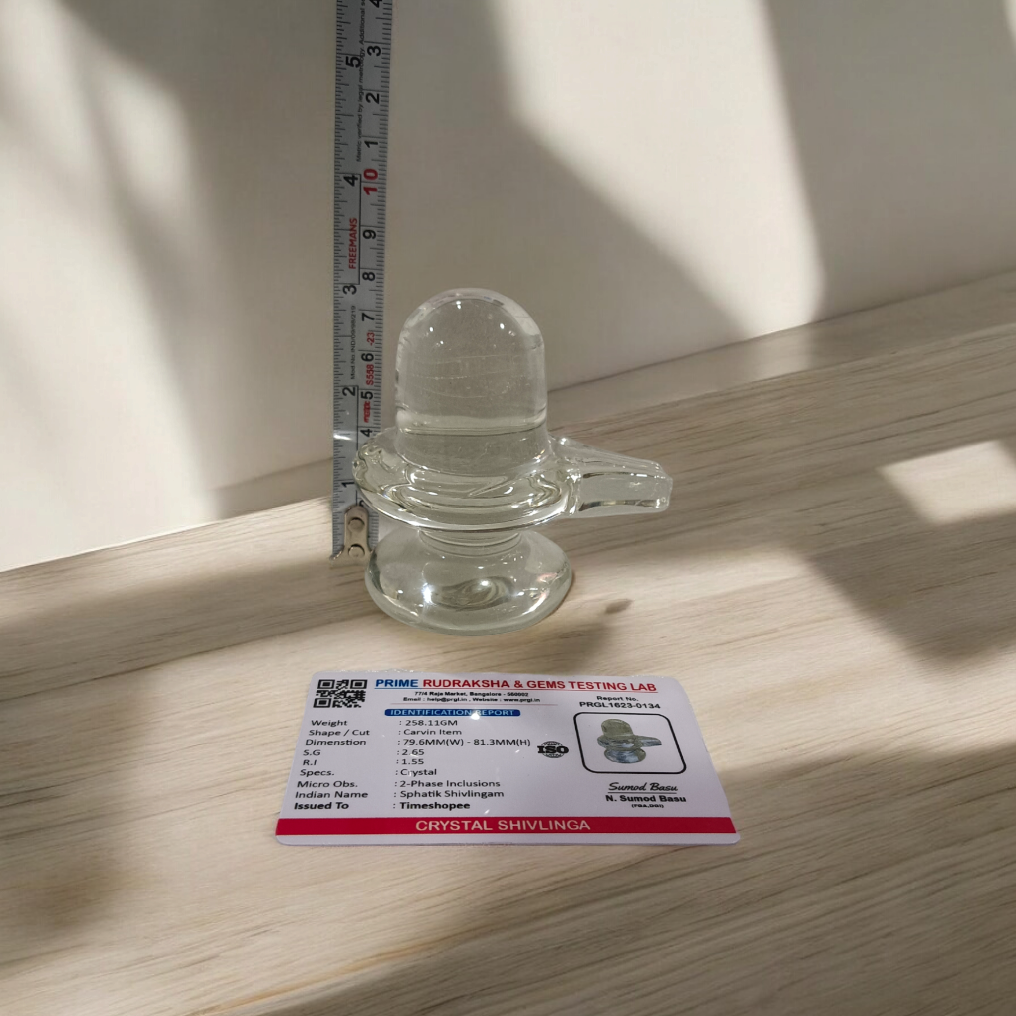 A clear, polished Sphatik (Crystal) Shivling placed on a small round base, intricately designed with fine details. The Shivling radiates purity and divine energy, symbolizing Lord Shiva. Ideal for worship, this lab-certified Sphatik Shivling from Timeshopee adds a spiritual touch to your home or temple setting.