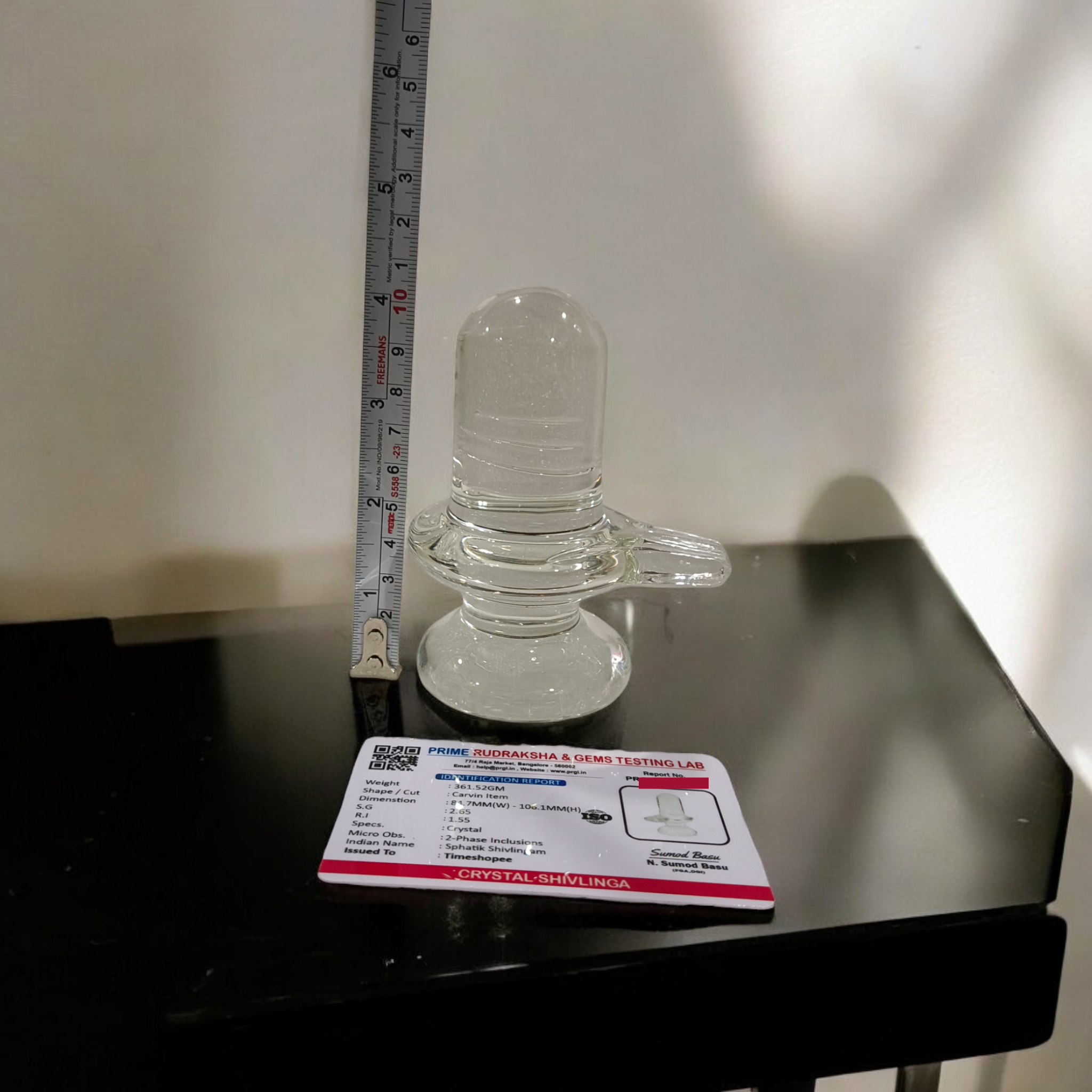 A clear, polished Sphatik (Crystal) Shivling placed on a small round base, intricately designed with fine details. The Shivling radiates purity and divine energy, symbolizing Lord Shiva. Ideal for worship, this lab-certified Sphatik Shivling from Timeshopee adds a spiritual touch to your home or temple setting.