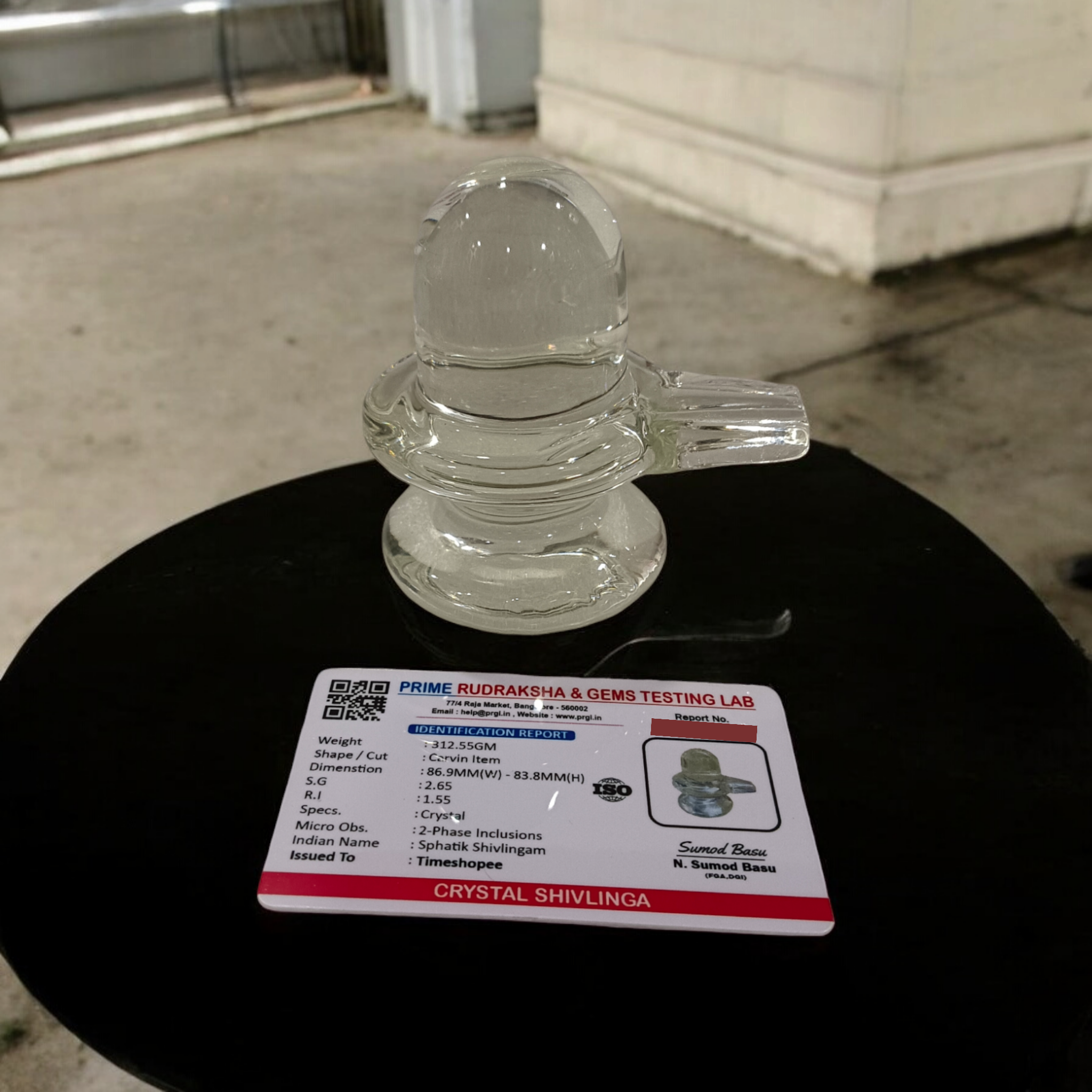 A clear, polished Sphatik (Crystal) Shivling placed on a small round base, intricately designed with fine details. The Shivling radiates purity and divine energy, symbolizing Lord Shiva. Ideal for worship, this lab-certified Sphatik Shivling from Timeshopee adds a spiritual touch to your home or temple setting.