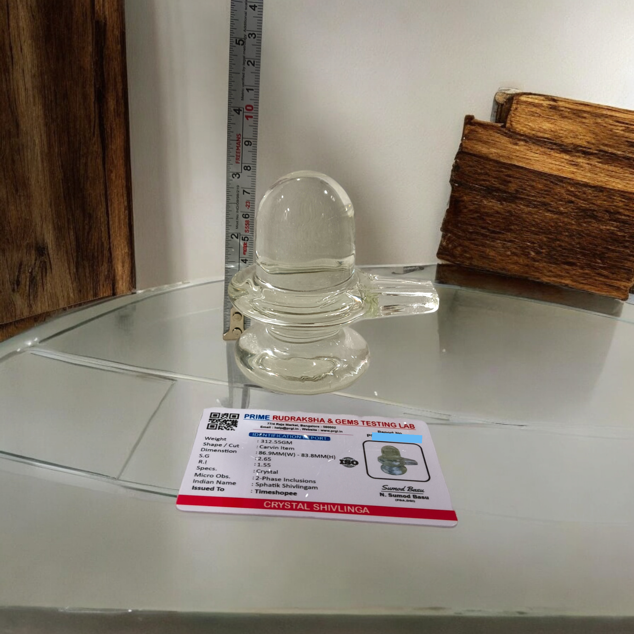 A clear, polished Sphatik (Crystal) Shivling placed on a small round base, intricately designed with fine details. The Shivling radiates purity and divine energy, symbolizing Lord Shiva. Ideal for worship, this lab-certified Sphatik Shivling from Timeshopee adds a spiritual touch to your home or temple setting.