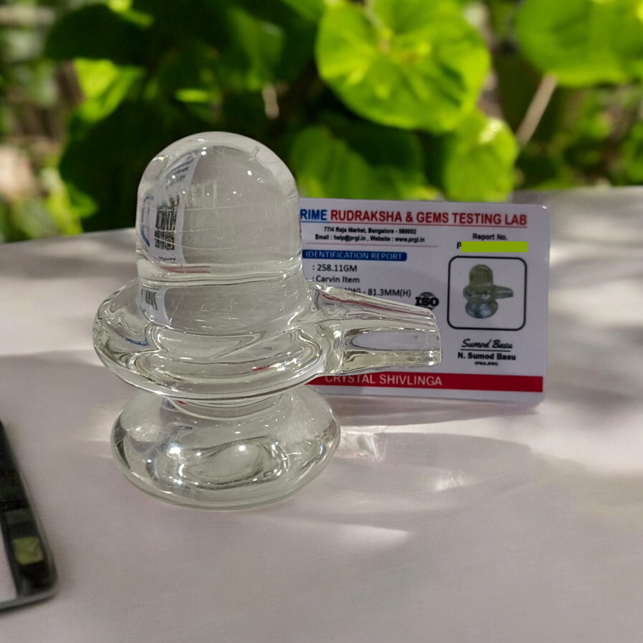 A clear, polished Sphatik (Crystal) Shivling placed on a small round base, intricately designed with fine details. The Shivling radiates purity and divine energy, symbolizing Lord Shiva. Ideal for worship, this lab-certified Sphatik Shivling from Timeshopee adds a spiritual touch to your home or temple setting.