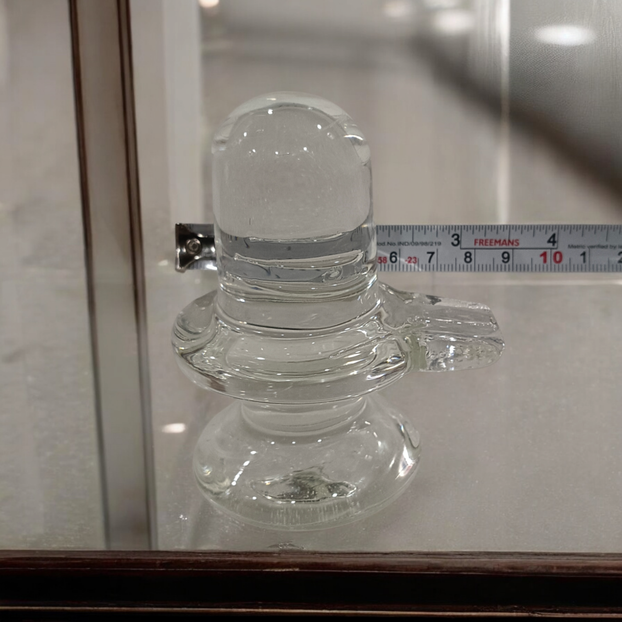 A clear, polished Sphatik (Crystal) Shivling placed on a small round base, intricately designed with fine details. The Shivling radiates purity and divine energy, symbolizing Lord Shiva. Ideal for worship, this lab-certified Sphatik Shivling from Timeshopee adds a spiritual touch to your home or temple setting.