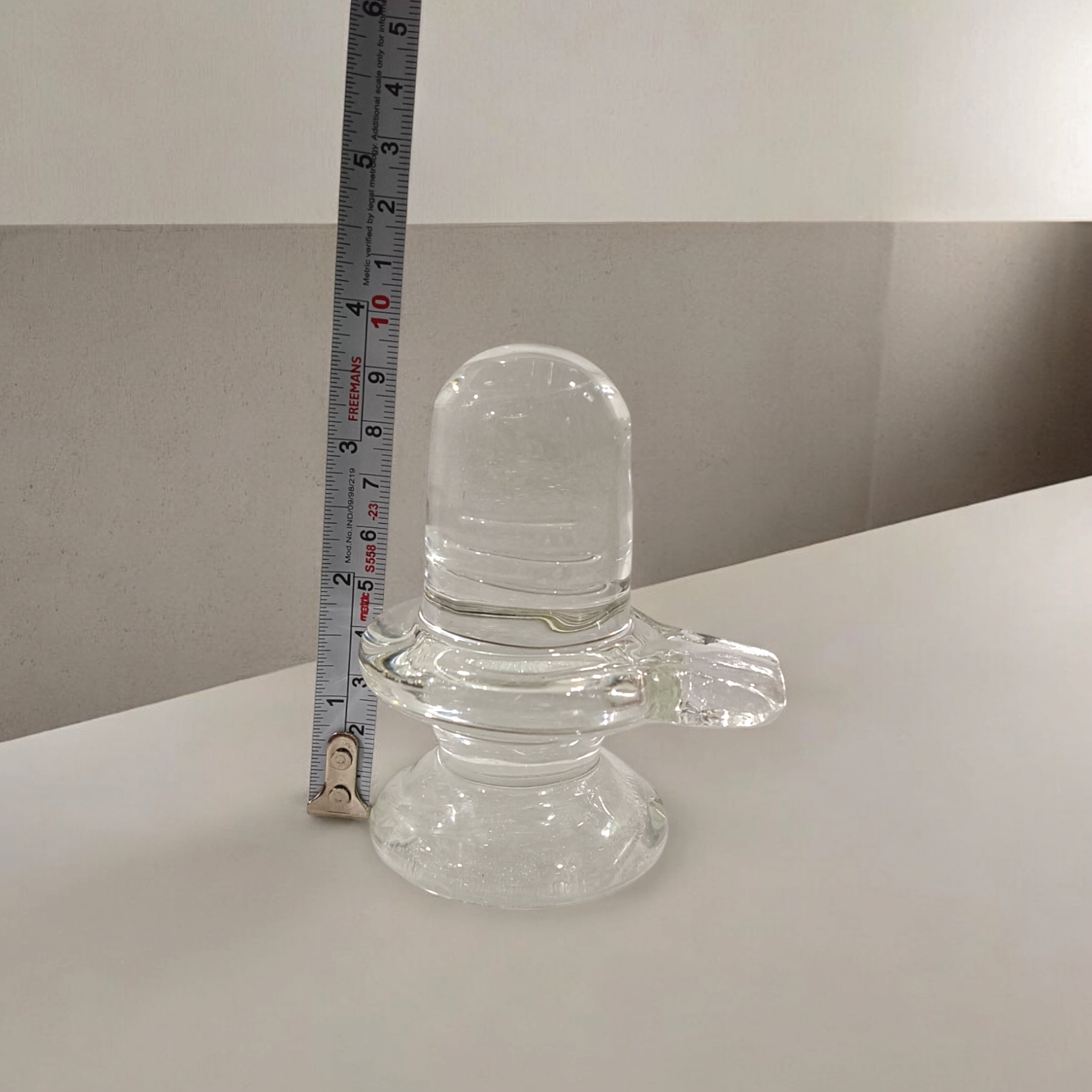 A clear, polished Sphatik (Crystal) Shivling placed on a small round base, intricately designed with fine details. The Shivling radiates purity and divine energy, symbolizing Lord Shiva. Ideal for worship, this lab-certified Sphatik Shivling from Timeshopee adds a spiritual touch to your home or temple setting.