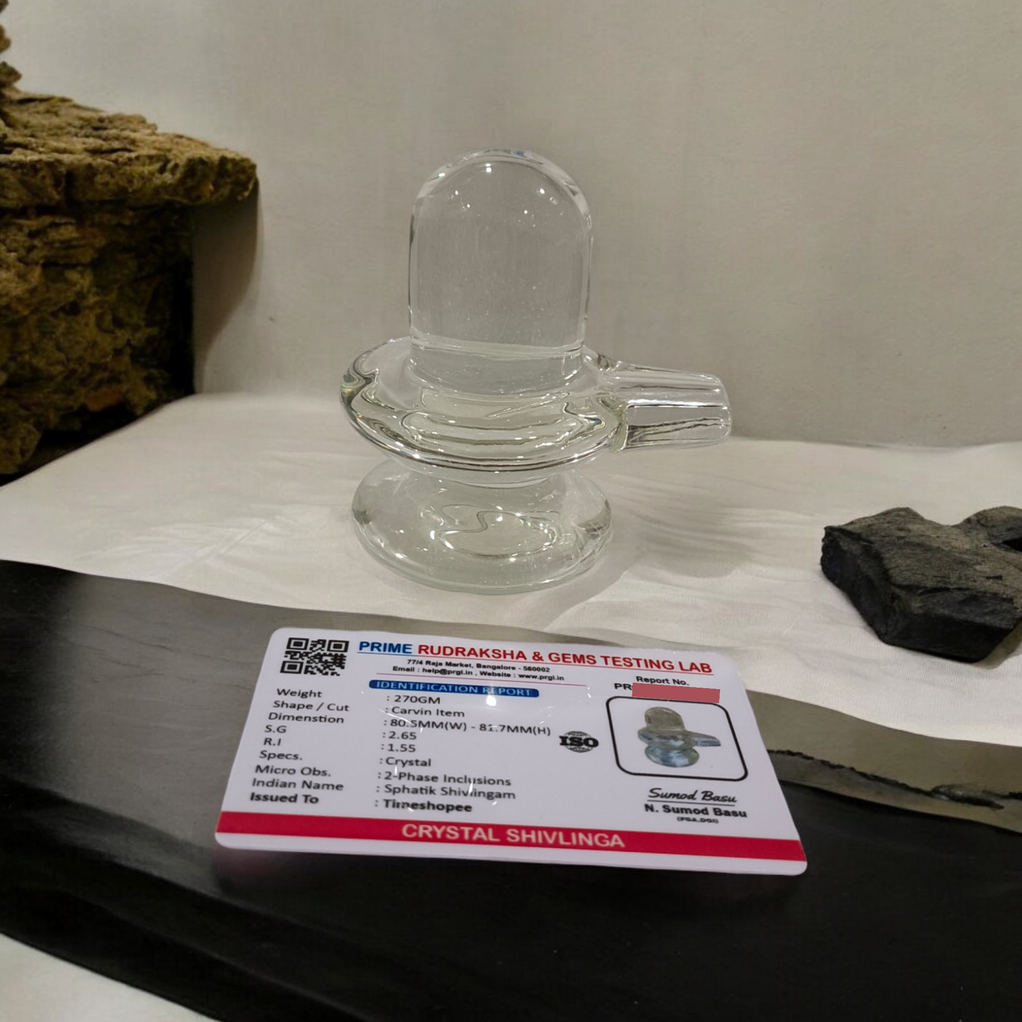 A clear, polished Sphatik (Crystal) Shivling placed on a small round base, intricately designed with fine details. The Shivling radiates purity and divine energy, symbolizing Lord Shiva. Ideal for worship, this lab-certified Sphatik Shivling from Timeshopee adds a spiritual touch to your home or temple setting.