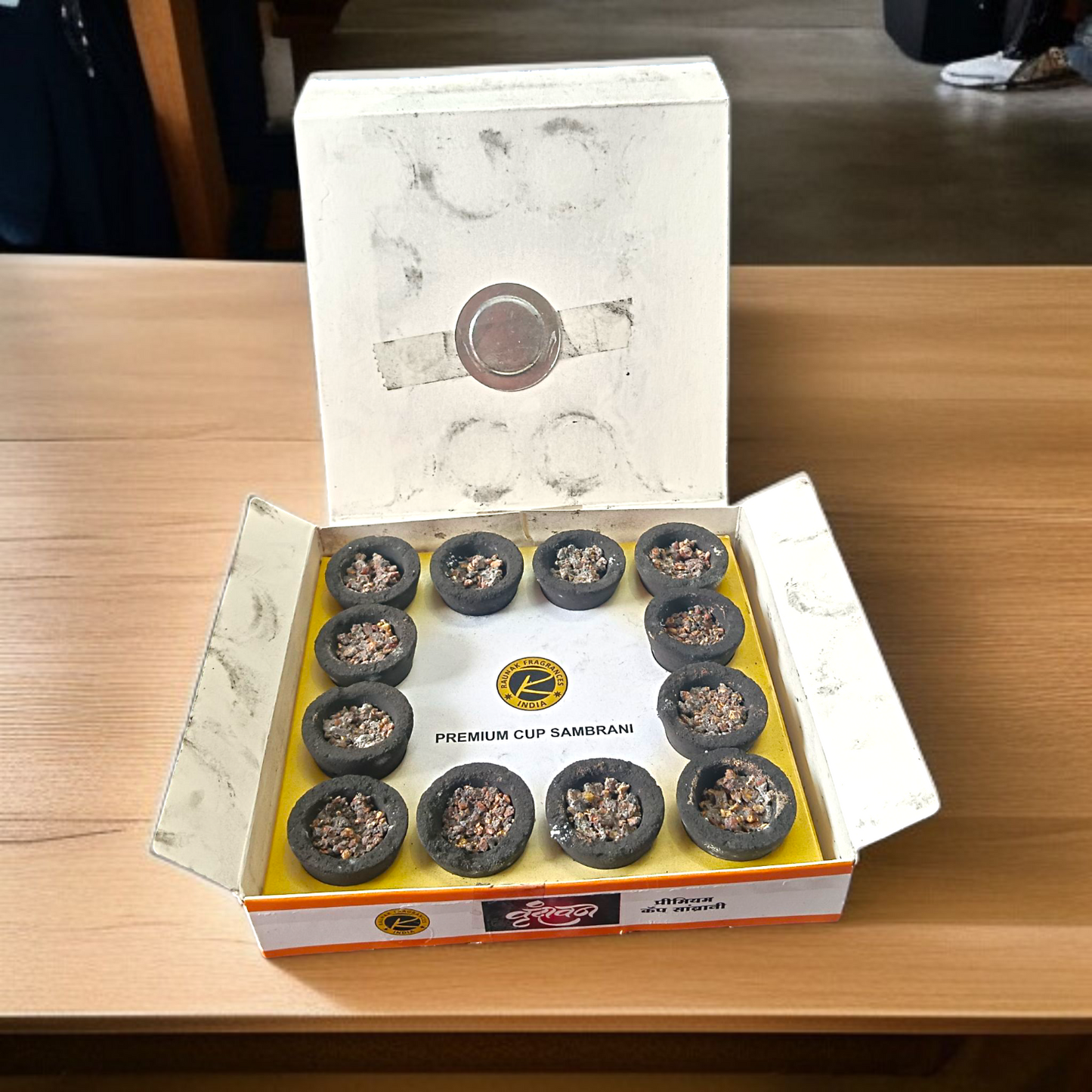 A box of Premium Cup Sambrani containing 12 natural dhoop cups arranged neatly, with herbal ingredients in the background