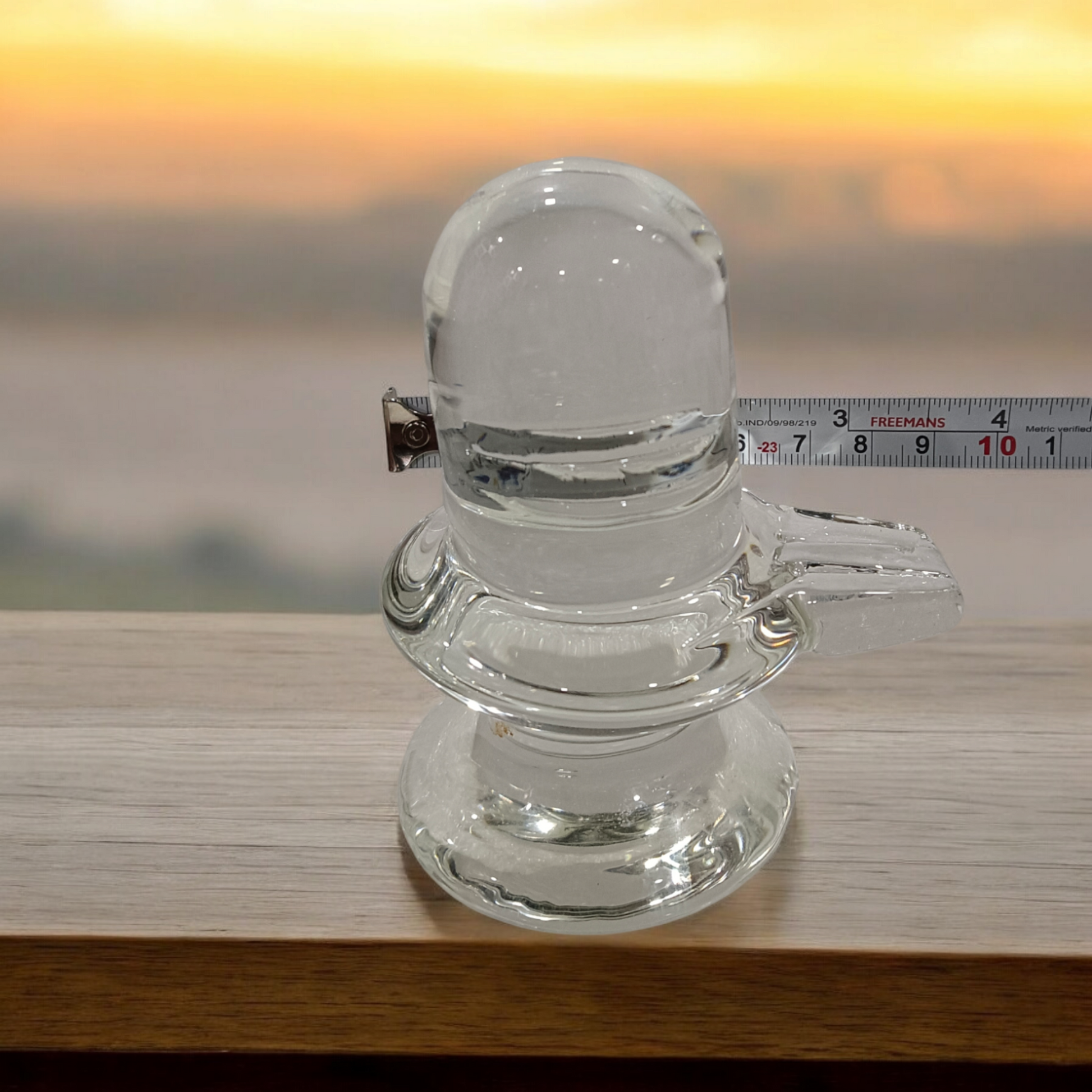 A clear, polished Sphatik (Crystal) Shivling placed on a small round base, intricately designed with fine details. The Shivling radiates purity and divine energy, symbolizing Lord Shiva. Ideal for worship, this lab-certified Sphatik Shivling from Timeshopee adds a spiritual touch to your home or temple setting.