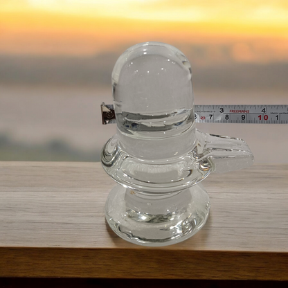 A clear, polished Sphatik (Crystal) Shivling placed on a small round base, intricately designed with fine details. The Shivling radiates purity and divine energy, symbolizing Lord Shiva. Ideal for worship, this lab-certified Sphatik Shivling from Timeshopee adds a spiritual touch to your home or temple setting.