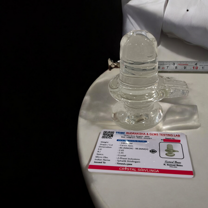 A clear, polished Sphatik (Crystal) Shivling placed on a small round base, intricately designed with fine details. The Shivling radiates purity and divine energy, symbolizing Lord Shiva. Ideal for worship, this lab-certified Sphatik Shivling from Timeshopee adds a spiritual touch to your home or temple setting.