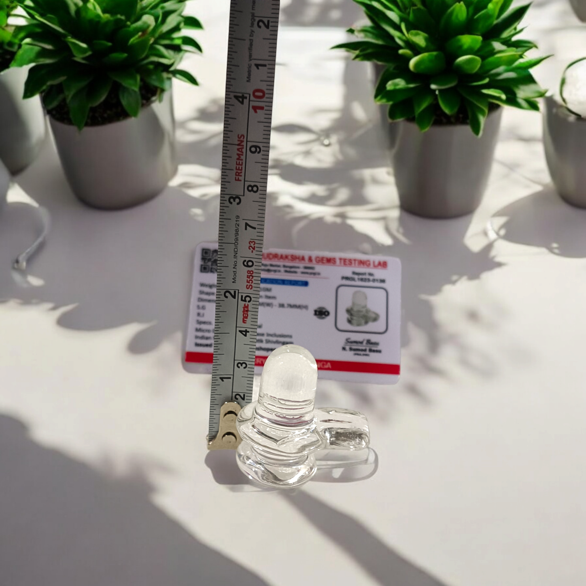 A clear, polished Sphatik (Crystal) Shivling placed on a small round base, intricately designed with fine details. The Shivling radiates purity and divine energy, symbolizing Lord Shiva. Ideal for worship, this lab-certified Sphatik Shivling from Timeshopee adds a spiritual touch to your home or temple setting.