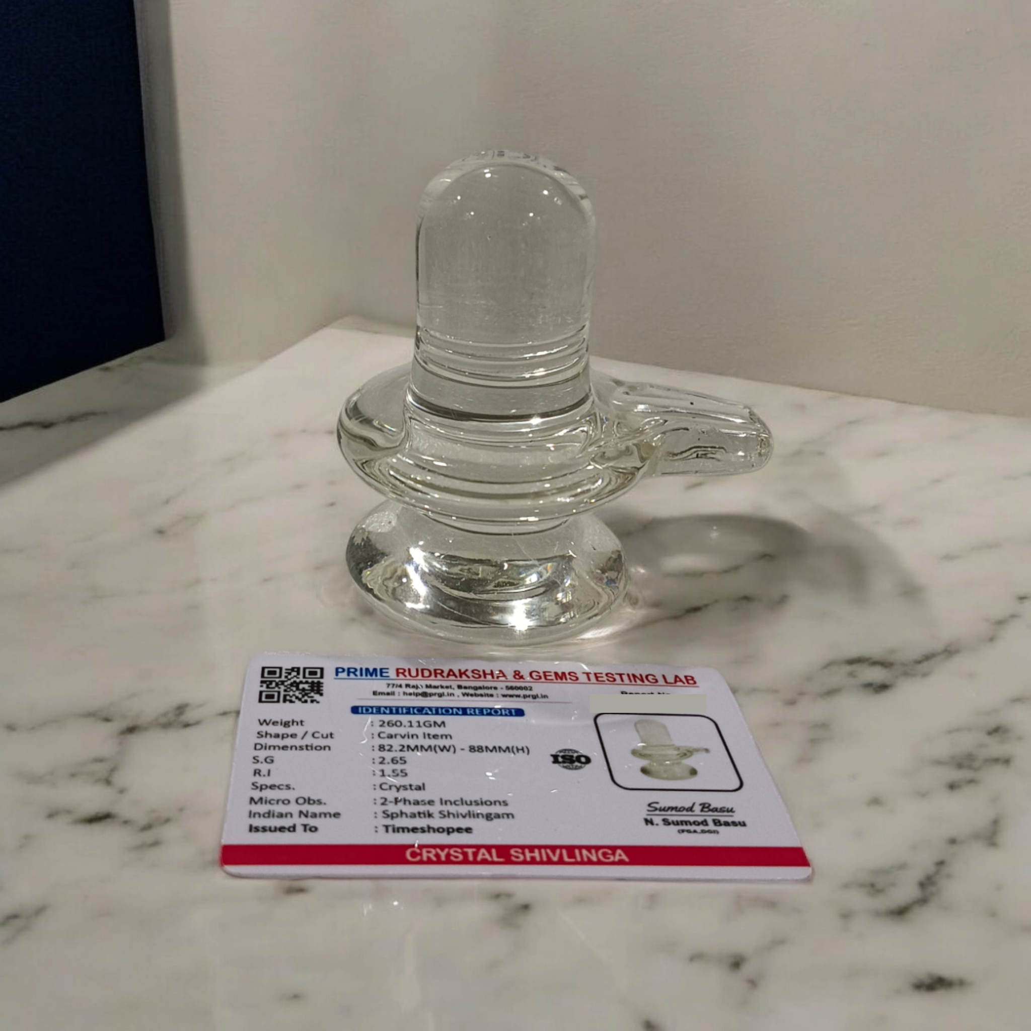 A clear, polished Sphatik (Crystal) Shivling placed on a small round base, intricately designed with fine details. The Shivling radiates purity and divine energy, symbolizing Lord Shiva. Ideal for worship, this lab-certified Sphatik Shivling from Timeshopee adds a spiritual touch to your home or temple setting.
