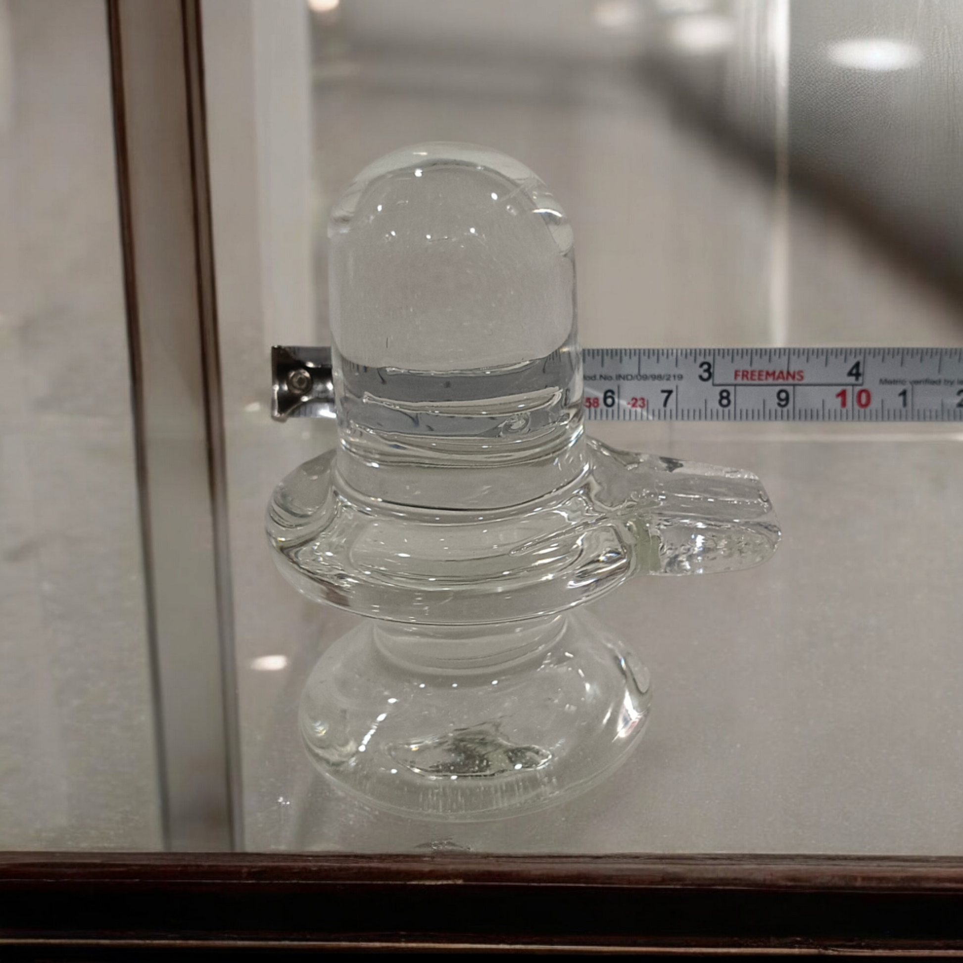A clear, polished Sphatik (Crystal) Shivling placed on a small round base, intricately designed with fine details. The Shivling radiates purity and divine energy, symbolizing Lord Shiva. Ideal for worship, this lab-certified Sphatik Shivling from Timeshopee adds a spiritual touch to your home or temple setting.